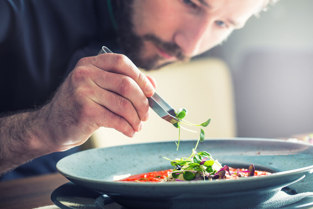 Chef in professional kitchen cooking, placing a micro herb on a tomato soup.