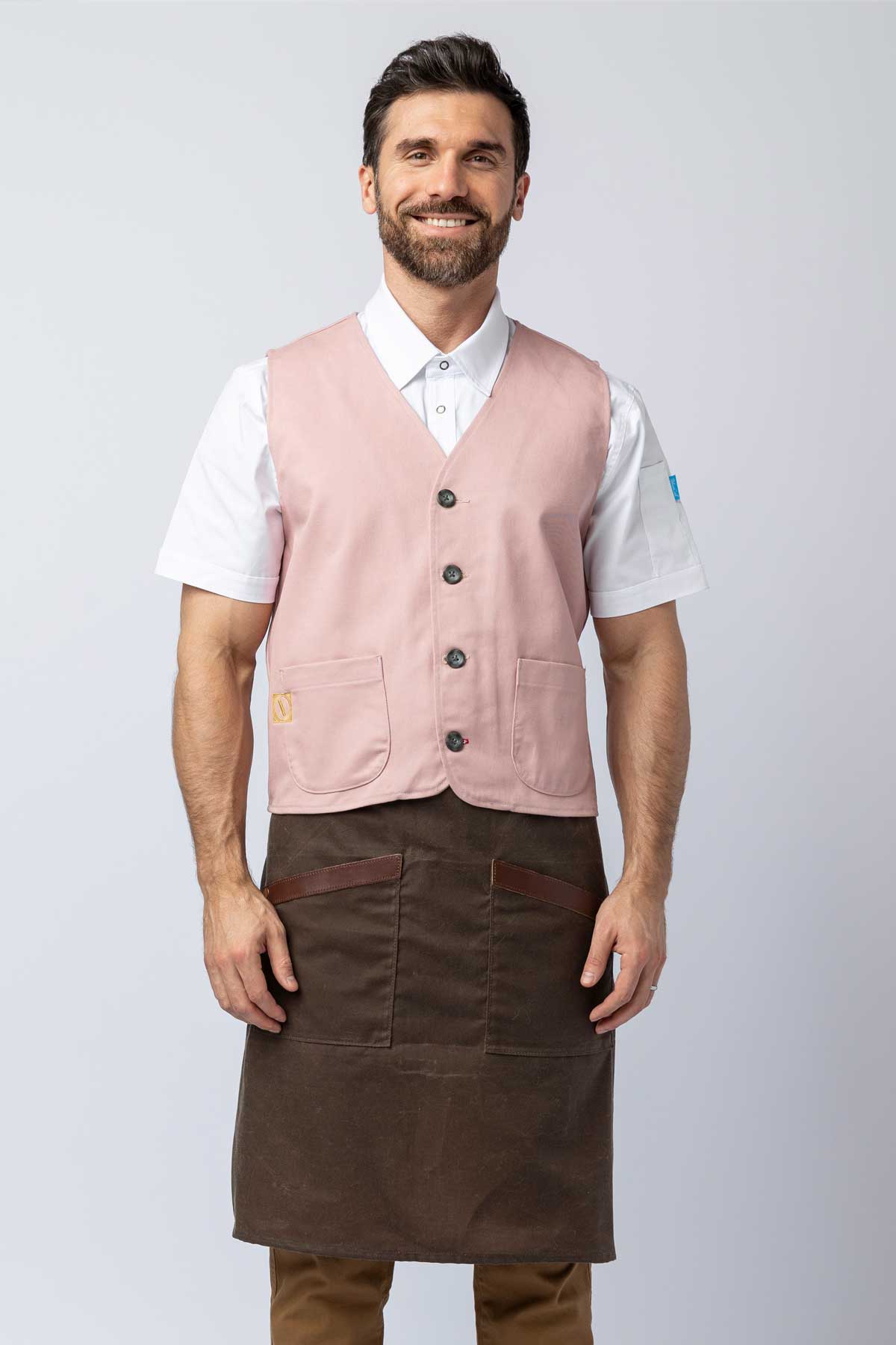 Man wearing a pink vest over a white shirt with a brown apron on a light gray background