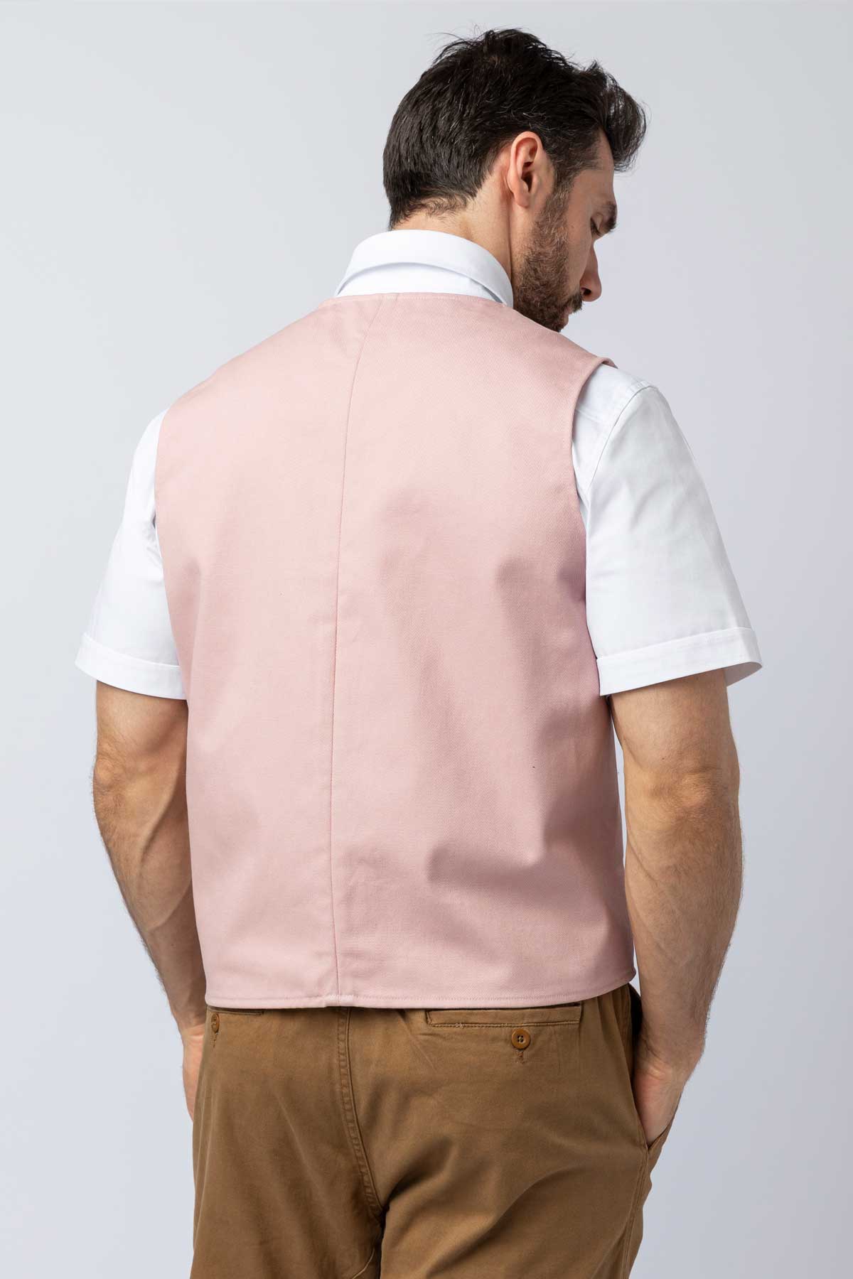Back view of man wearing a light pink vest over a white shirt with brown pants on a plain background