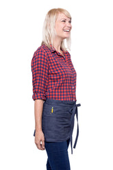 A side view of person wearing a indigo mini denim apron with a waist tie and pockets, standing against a neutral background.