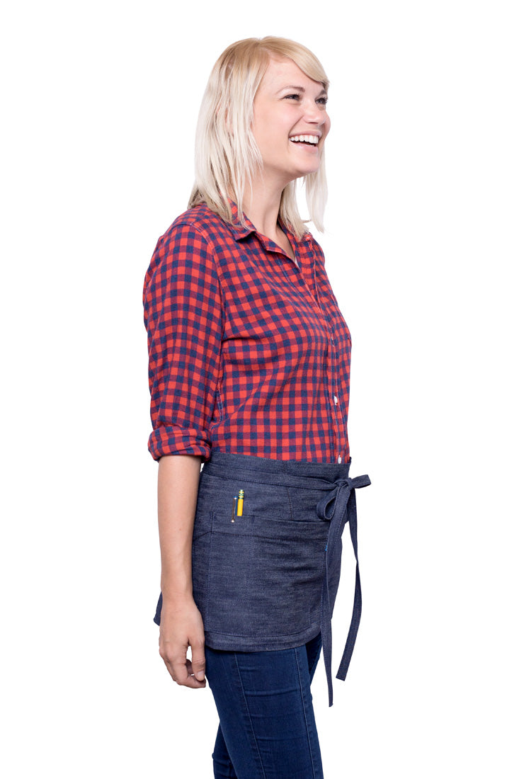 A side view of person wearing a indigo mini denim apron with a waist tie and pockets, standing against a neutral background.