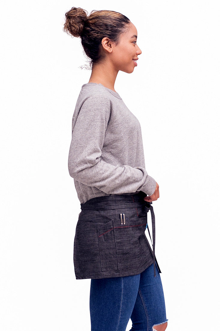 A side view of person wearing a charcoal mini denim apron with a waist tie and pockets, standing against a neutral background.
