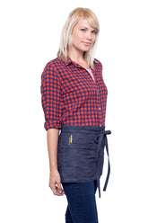 A side view of person wearing a indigo mini denim apron with a waist tie and pockets, standing against a neutral background.