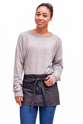 A person wearing a charcoal mini denim apron with a waist tie and pockets, standing against a neutral background.