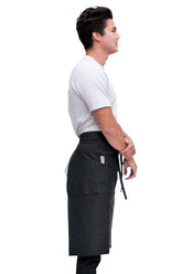 A profile view of a man wearing a black bistro apron with pockets and a tie at the waist, standing in a relaxed pose.