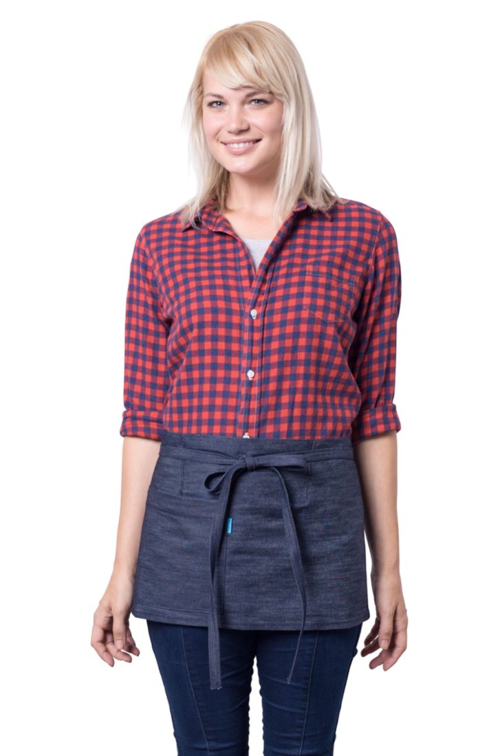 A person wearing a indigo mini denim apron with a waist tie and pockets, standing against a neutral background.