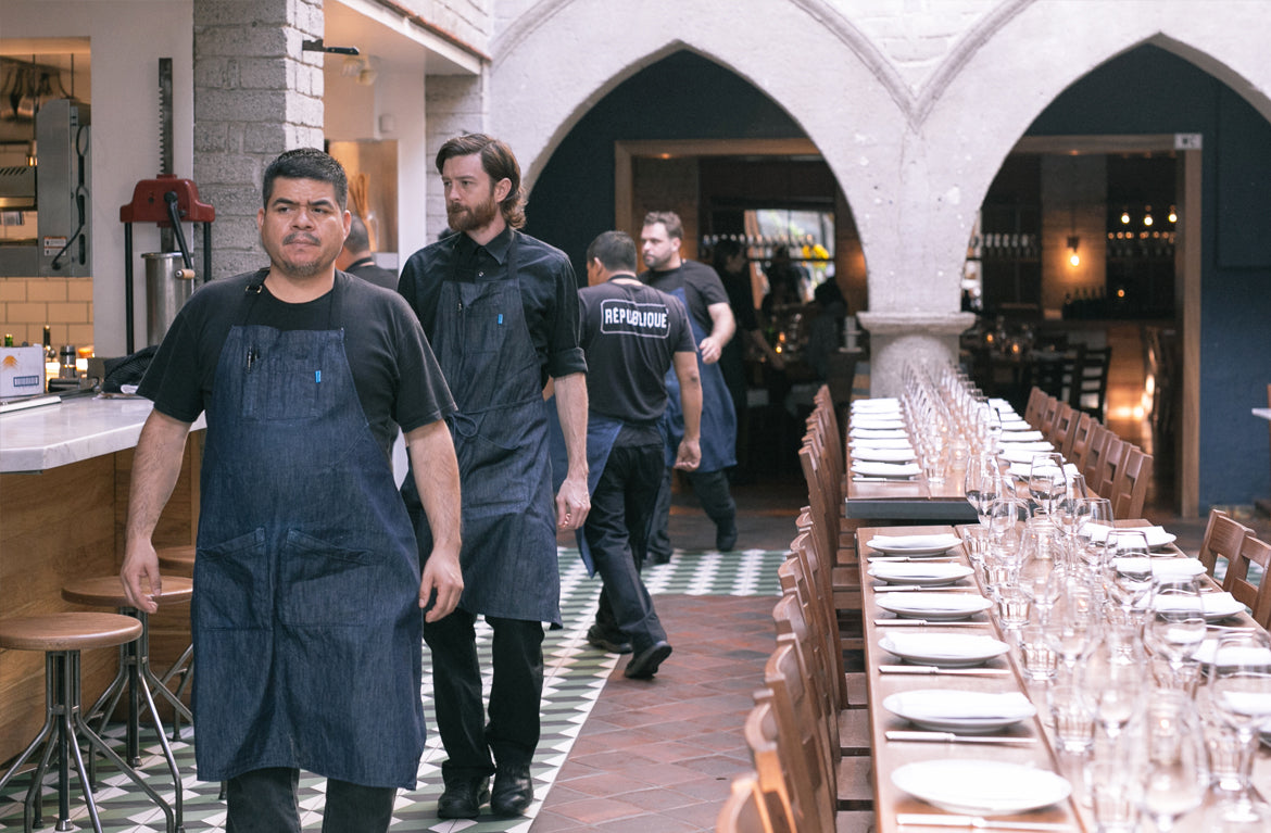 Restaurant staff wearing coordinated custom aprons walking through a branded dining space with long table setup