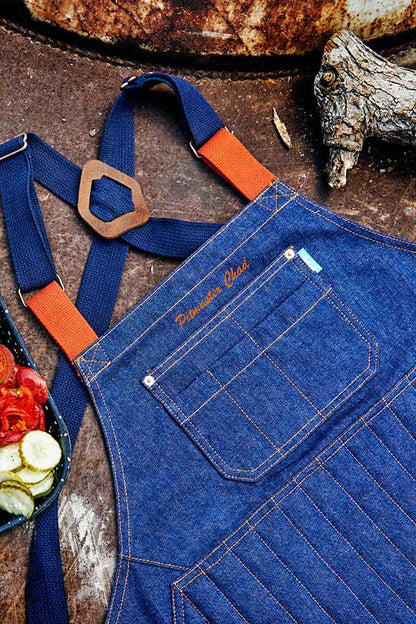 Blue denim Pitmaster Apron with orange name embroidery on a rustic background