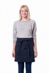 A smiling blonde woman wearing a light gray long-sleeve sweater, dark blue jeans, and a navy blue waist apron tied at the waist, standing against a plain white background.