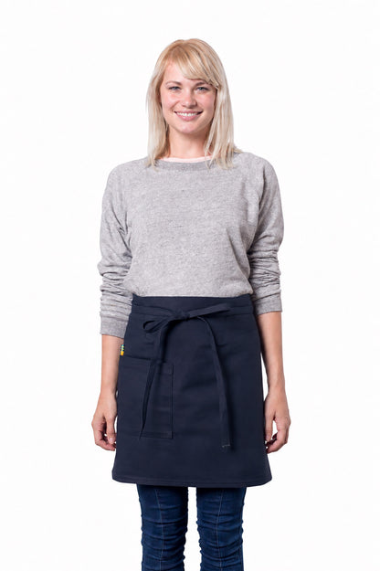 A smiling blonde woman wearing a light gray long-sleeve sweater, dark blue jeans, and a navy blue waist apron tied at the waist, standing against a plain white background.