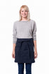 A smiling blonde woman wearing a light gray long-sleeve sweater, dark blue jeans, and a navy blue waist apron tied at the waist, standing against a plain white background.