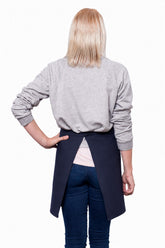 A blonde woman shown from the back, wearing a light gray long-sleeve sweater, dark blue jeans, and a navy blue waist apron tied at the waist, standing against a plain white background.