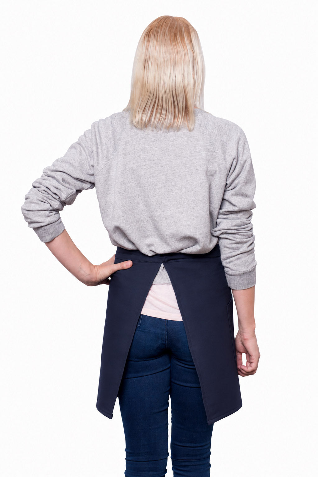 A blonde woman shown from the back, wearing a light gray long-sleeve sweater, dark blue jeans, and a navy blue waist apron tied at the waist, standing against a plain white background.